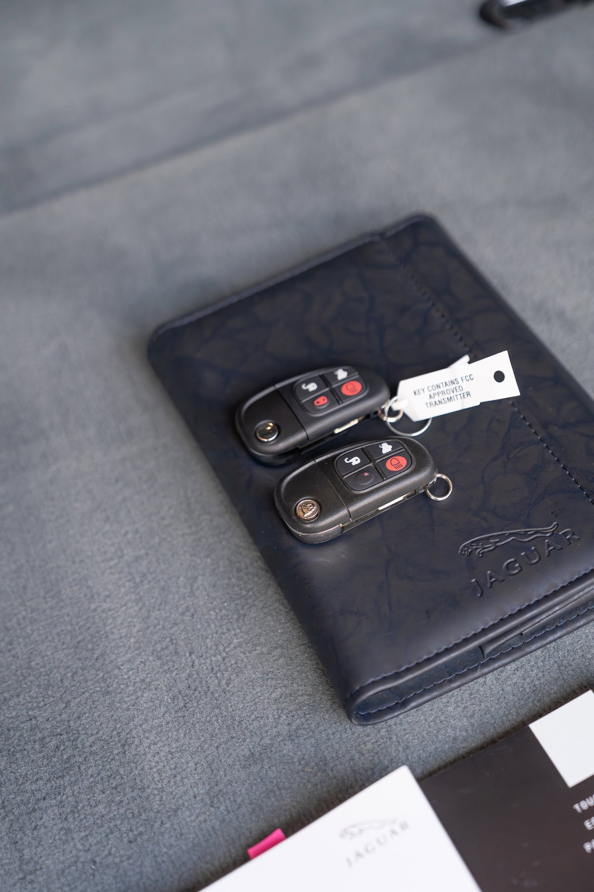 Two Jaguar XJR key fobs rest on a navy leather owner's manual. Radiance Red 2006 XJR.