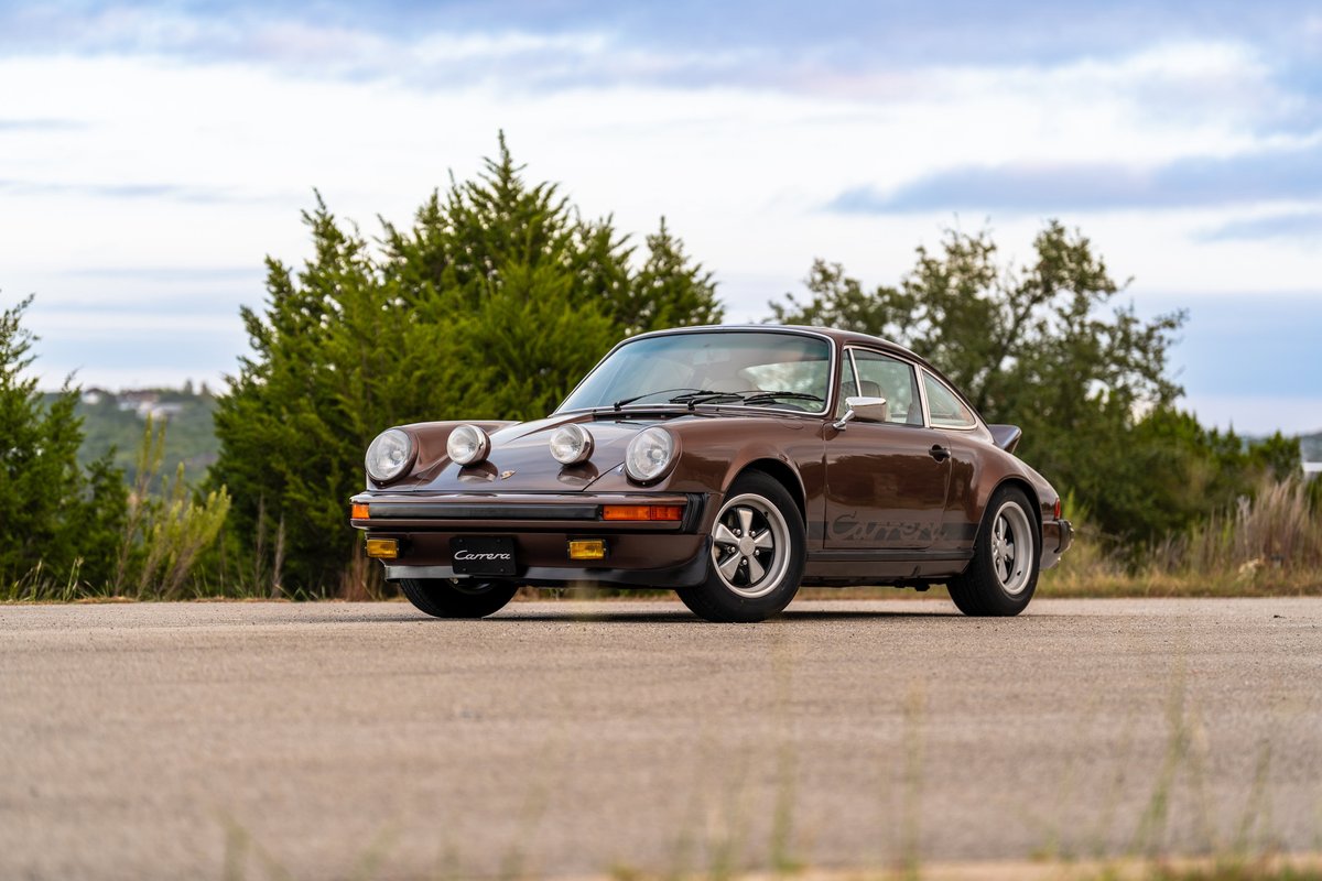 Brown metallic 1975 Porsche 911 Carrera, front view, on road with trees.