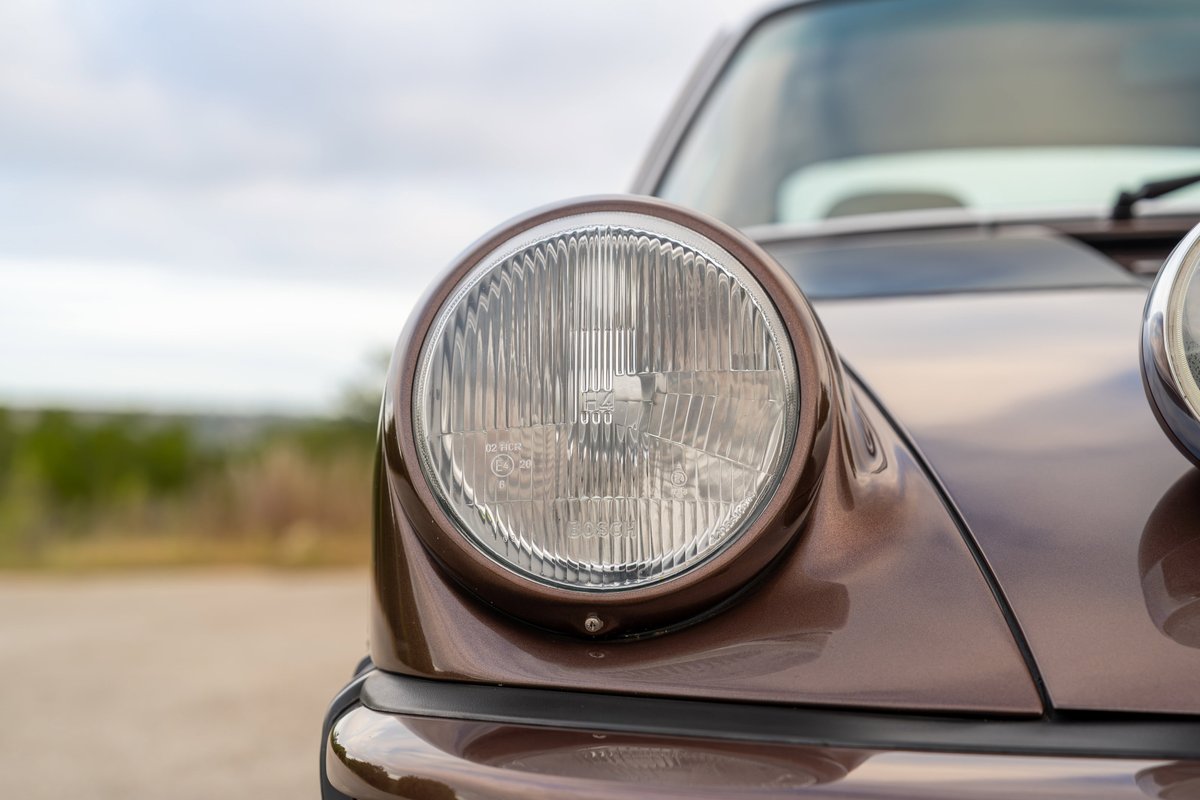 Close-up of the iconic round headlight on a copper brown metallic 1975 Porsche 911 Carrera.