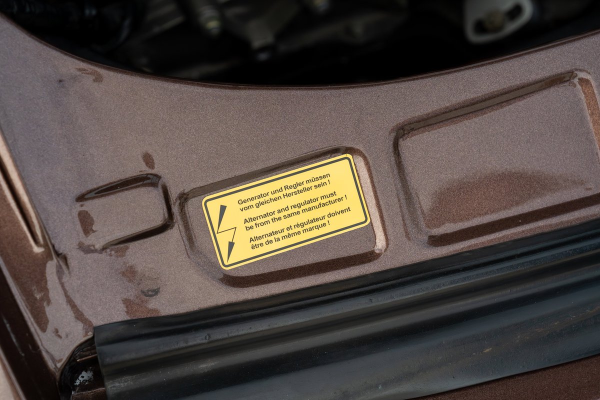 Close-up of a warning sticker on the brown metallic 1975 Porsche 911 Carrera engine bay.
