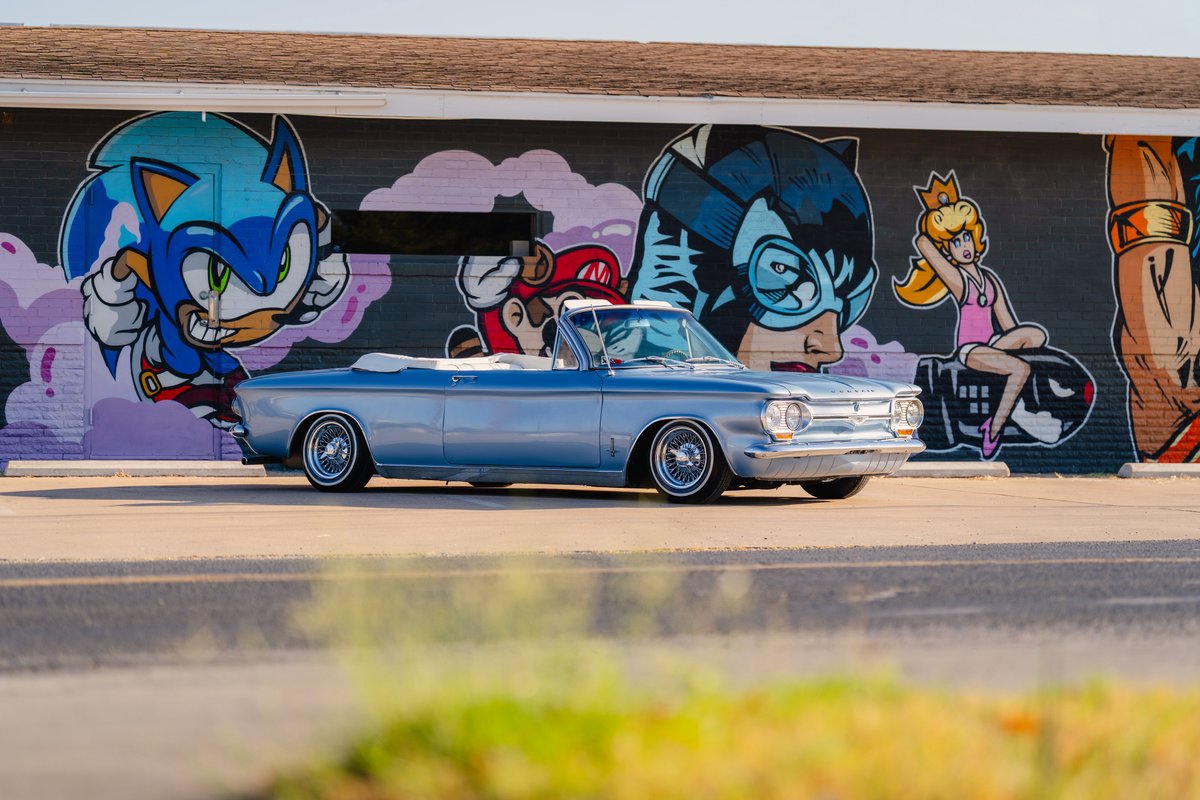 Blue metallic 1966 Chevrolet Corvair convertible with chrome rims and white wall tires parked in front of comic graffiti.