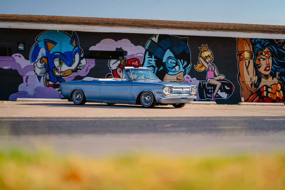 A blue metallic 1966 Chevrolet Corvair convertible with chrome wire wheels parked in front of a mural.