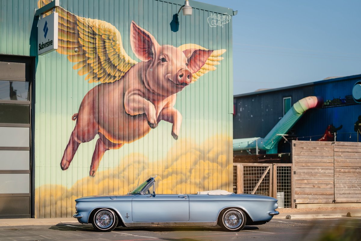 A metallic blue 1966 Chevrolet Corvair convertible with chrome rims sits parked in front of a mural.