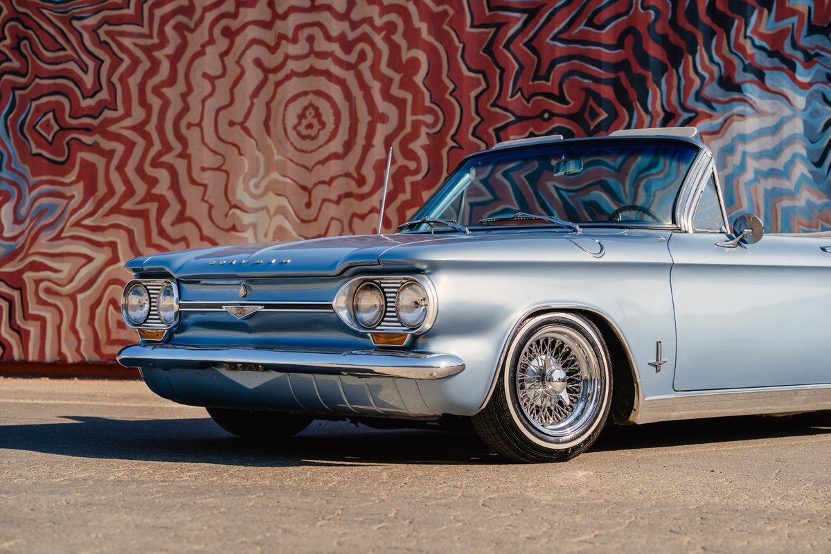 Blue 1966 Chevrolet Corvair convertible with chrome wire rims and whitewall tires parked by a mural.