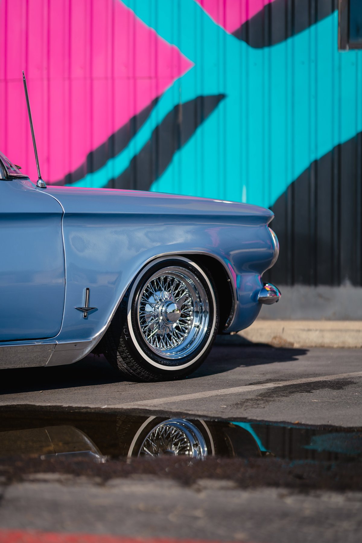 Blue 1966 Corvair convertible with chrome wire wheels and whitewall tires parked in front of a mural.