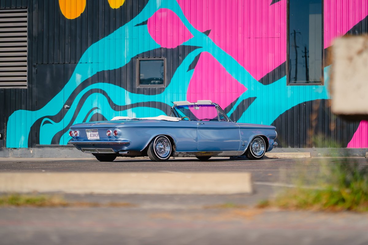 Blue 1966 Chevrolet Corvair convertible with chrome rims and white-wall tires parked in front of a colorful mural.