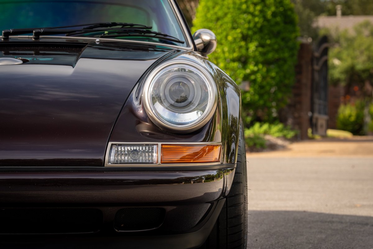 Close-up of a dark purple 1990 Singer Porsche 911, showing its iconic round headlight and amber turn signal.