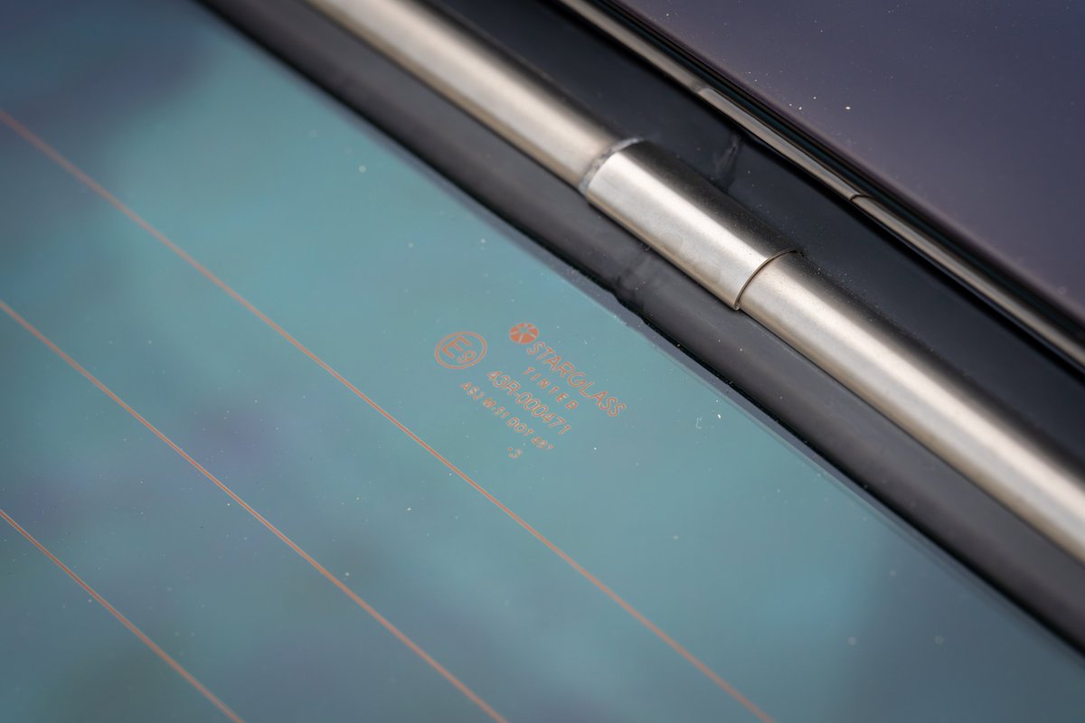 Close up of a 1990 Singer Porsche 911's rear windshield, showing heating elements and a branded manufacturer's mark.
