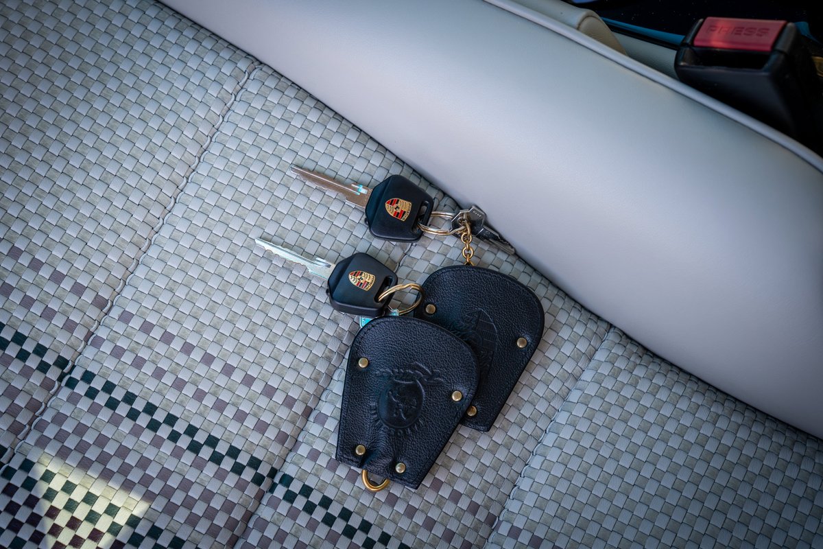Two Porsche 911 keys with leather fobs on aubergine metallic fabric seats.