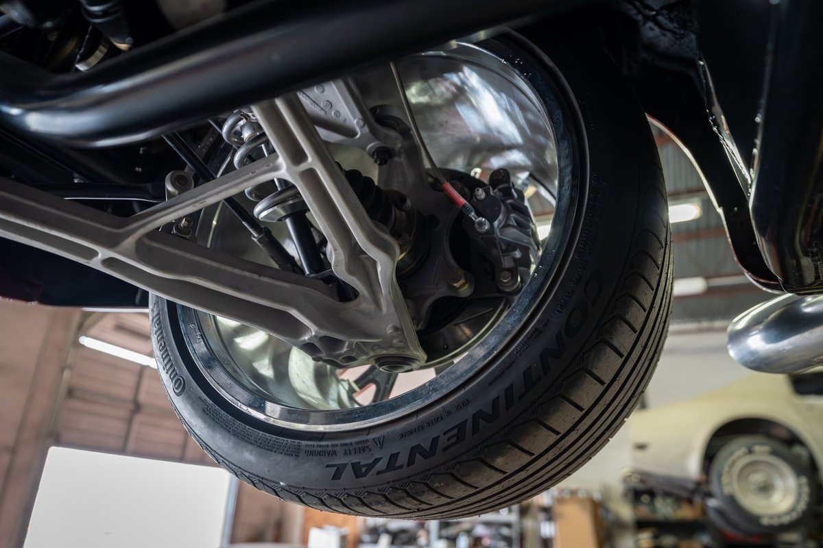 Close-up of a 1965 Chevrolet Corvette's rear suspension, showing alloy control arms, coilover shock, and Continental tire on polished rim.