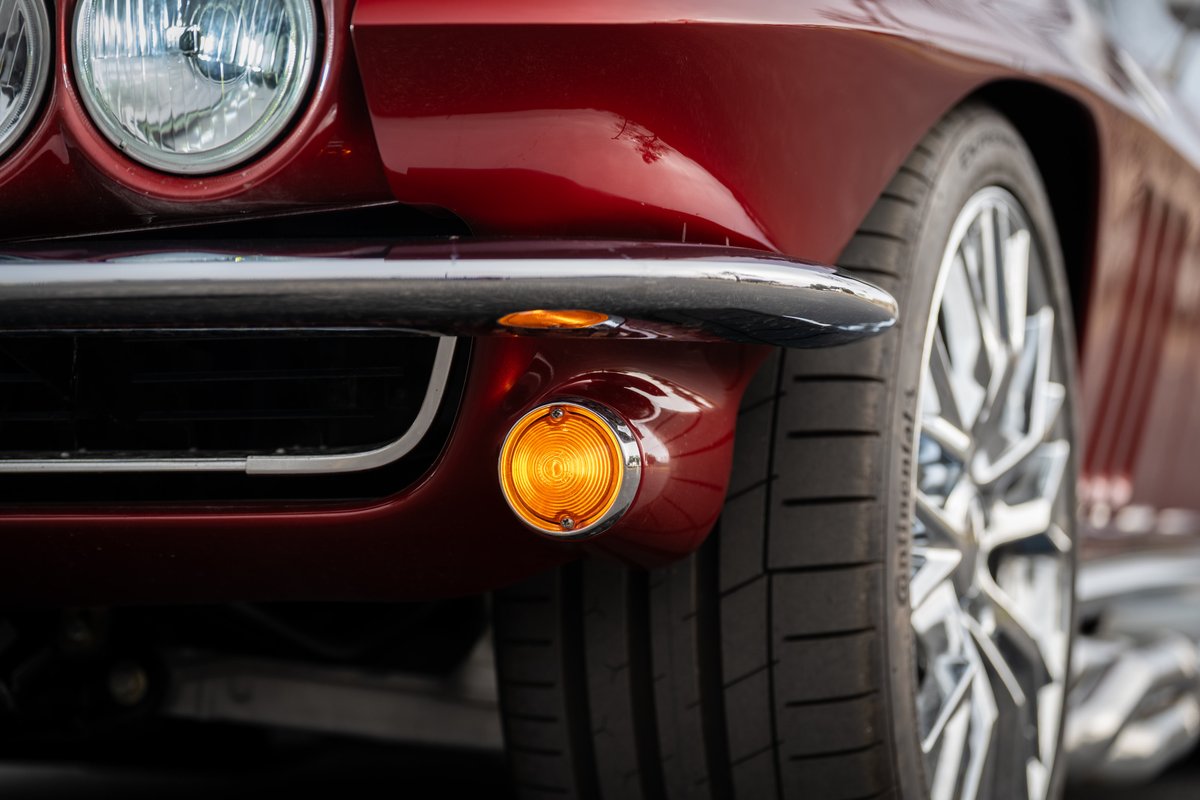 Close-up of a red 1965 Chevrolet Corvette's front bumper, chrome trim, and amber turn signal.