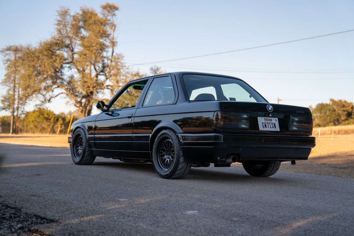 Rear view of a black 1989 BMW 325is with black mesh rims and tinted taillights.