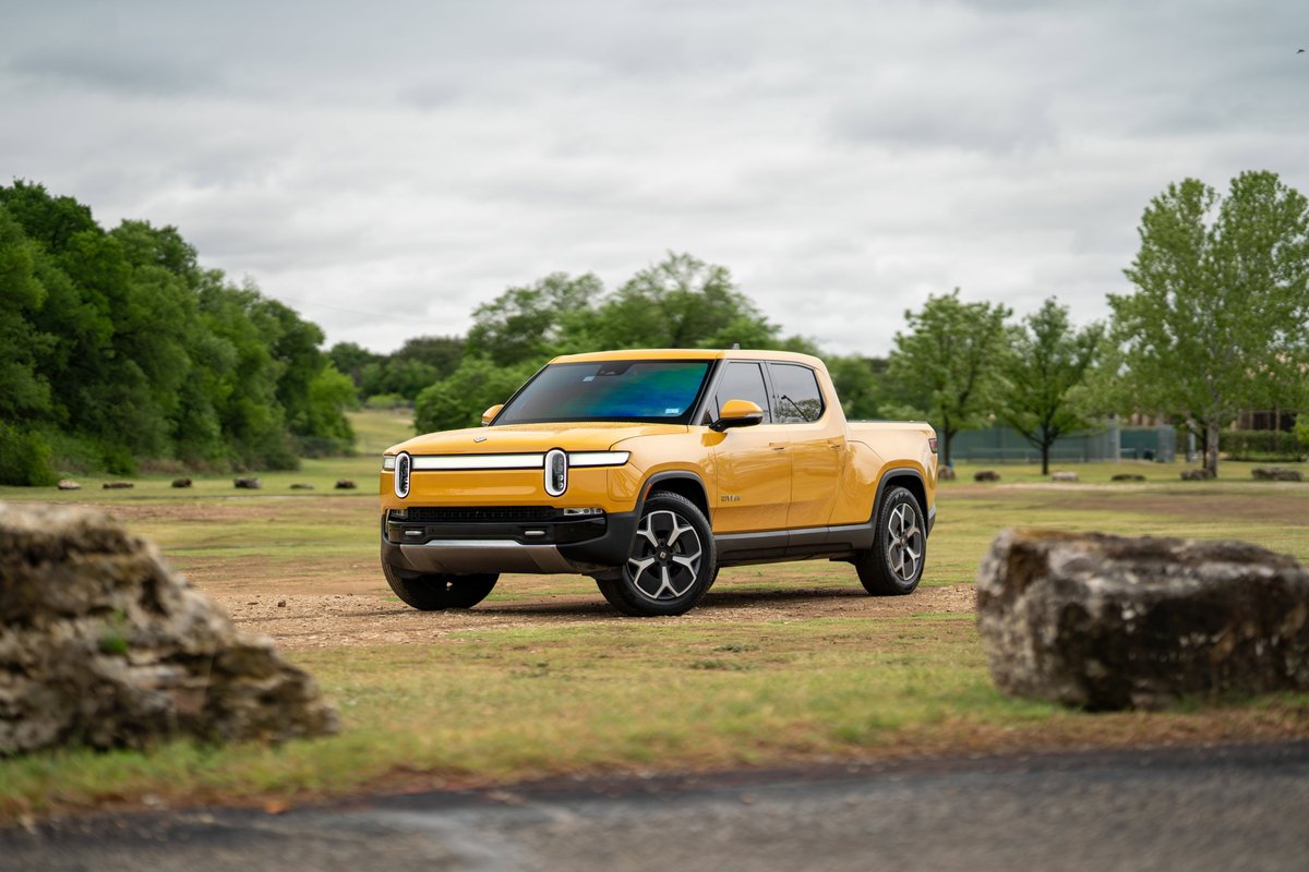 Yellow Rivian R1T electric pickup truck with black rims, parked in a field.