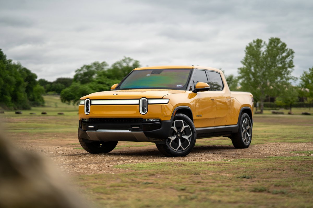 Yellow 2022 Rivian R1T pickup truck with distinctive rims and LED light bar, parked on dirt and grass.