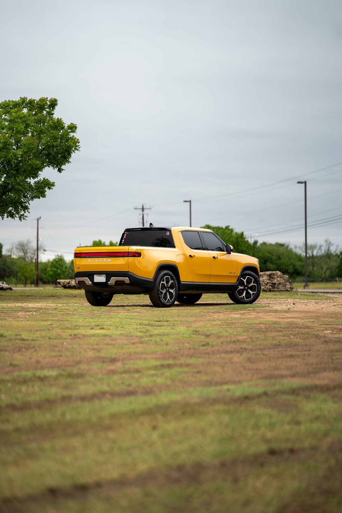 Yellow 2022 Rivian R1T pickup truck with black trim and sport rims parked on grass.