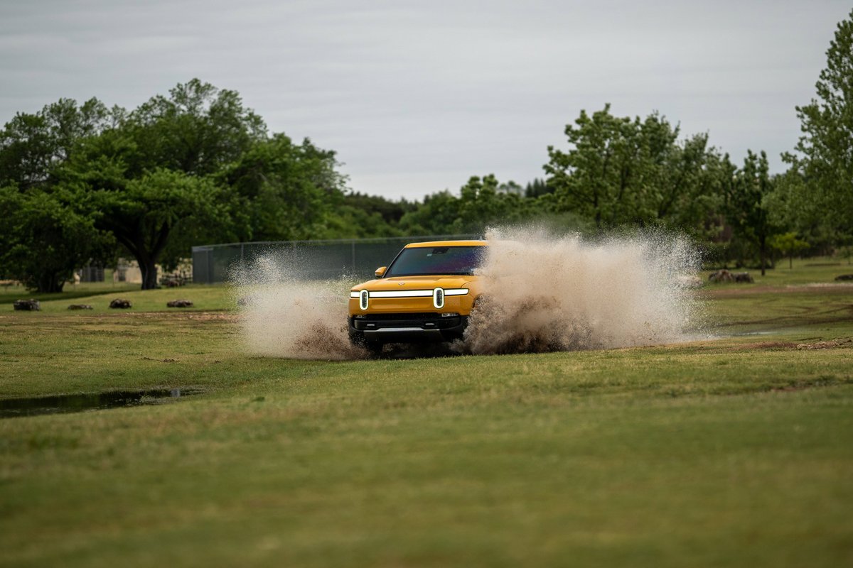 Yellow Rivian R1T SUV kicking up mud and water off-road.