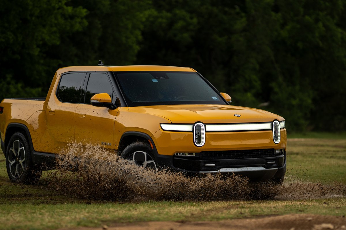 Yellow Rivian R1T electric truck driving through mud, featuring distinctive LED light bar and vertical headlights.
