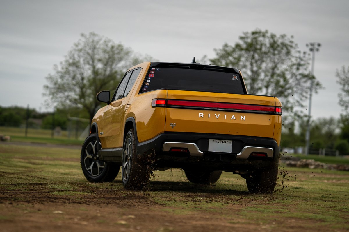 Rear view of a yellow Rivian R1T on dirt roads with custom rims and black trim.