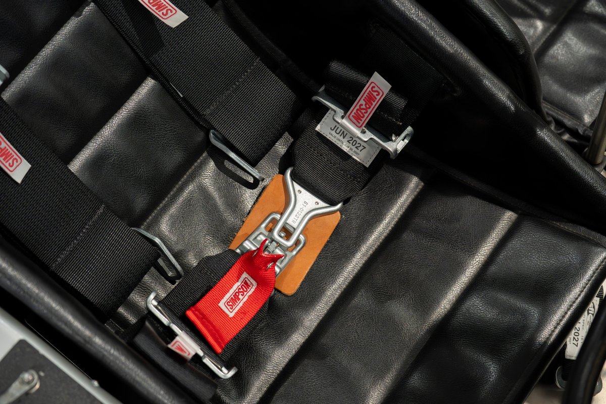 Close-up of a black leather racing seat with Simpson five-point harnesses, featuring red and black webbing.