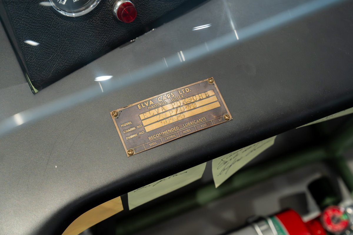 Close-up of a brass Elva Porsche 4-Cam chassis plate on a gray dashboard.