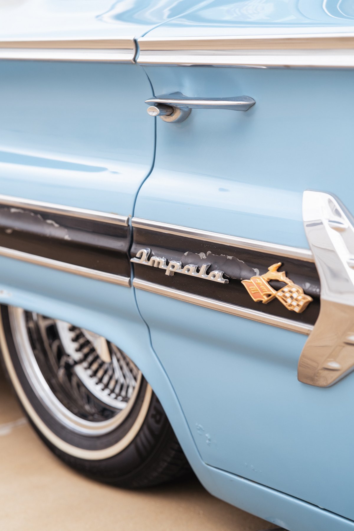 Close-up of a light blue 1963 Chevrolet Impala's rear fender with 'Impala' trim, gold checkered flag emblem, and wire wheels.