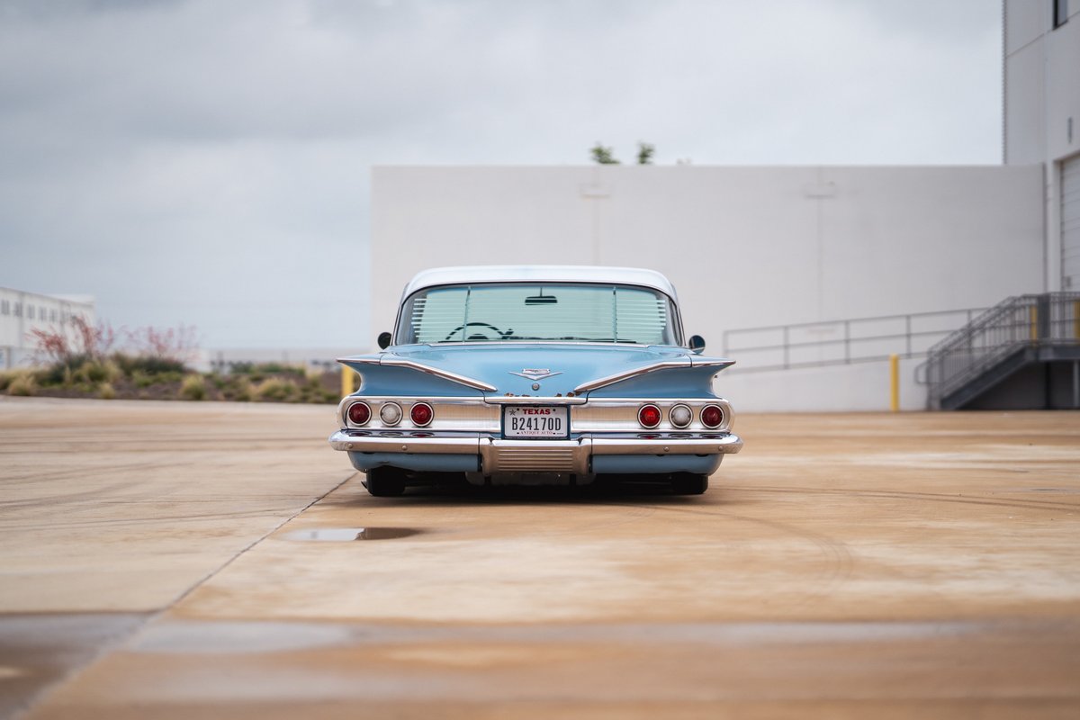 Rear view of a light blue 1960 Chevrolet Impala lowrider with prominent tail fins and chrome bumper.