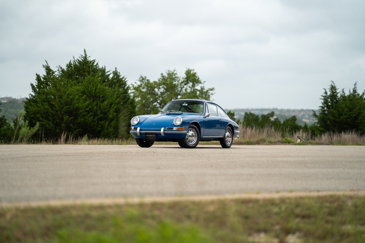 A blue 1965 Porsche 912 coupe sits parked on an asphalt road, with chrome rims and trim.