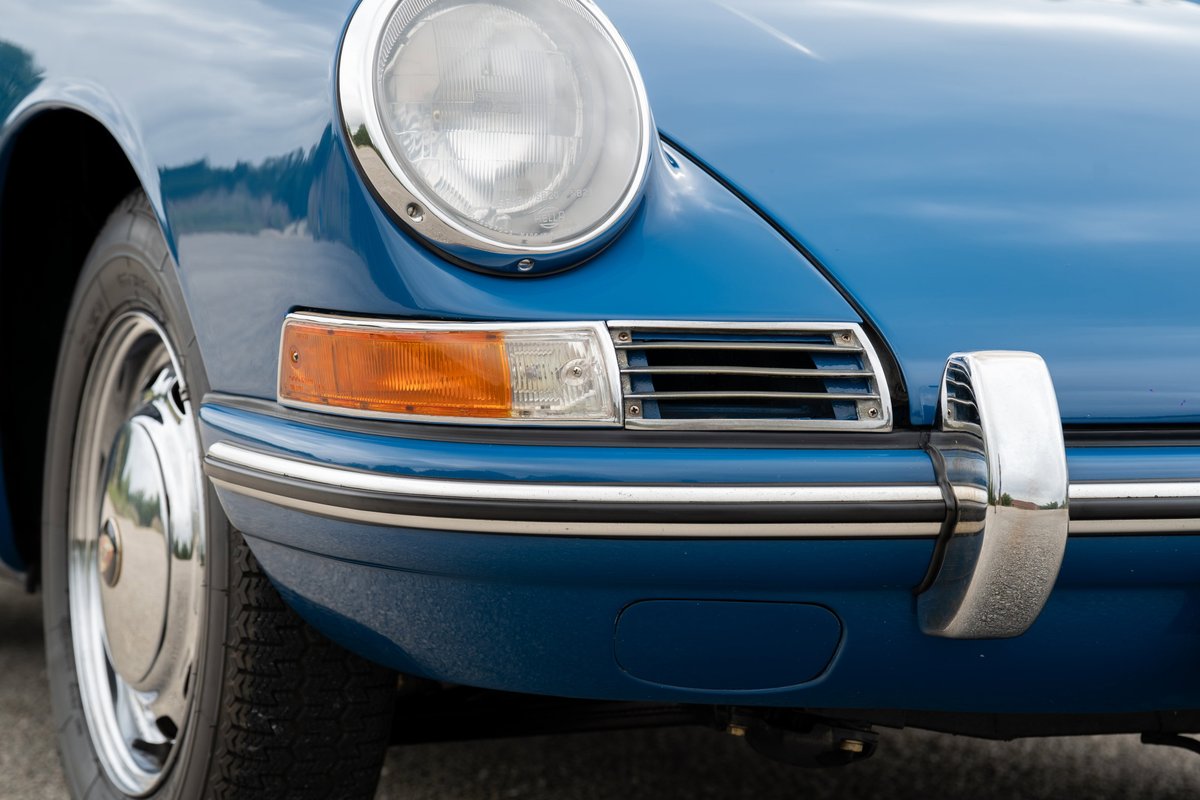 Close up of a blue 1965 Porsche 912 front bumper and headlight with chrome trim.