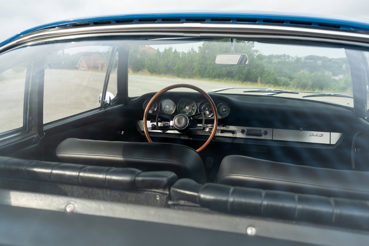 Interior of a blue 1965 Porsche 912, showing black leather seats, a wood-rimmed steering wheel, and silver dash trim.