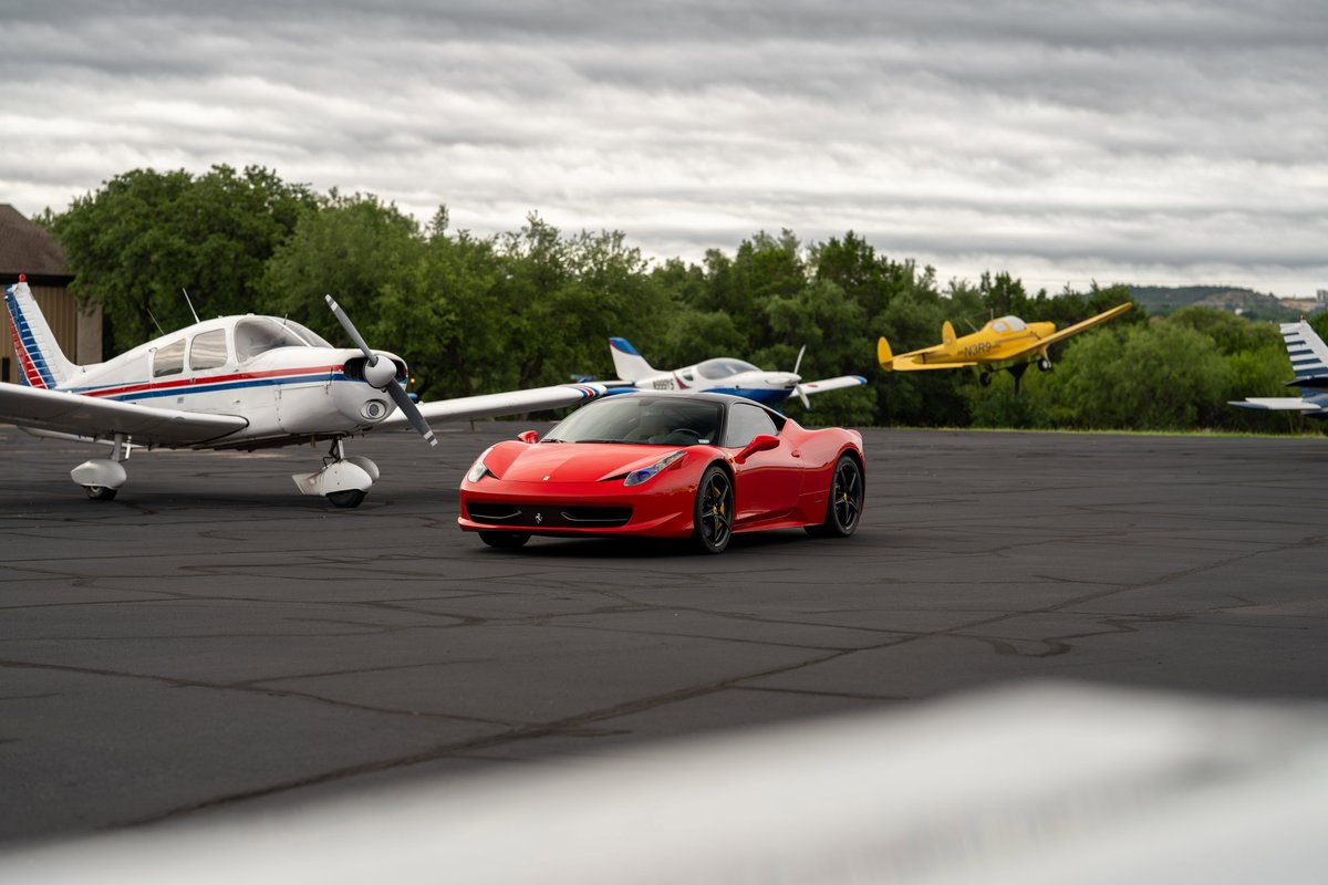 A red Ferrari 458 Italia with black rims parked on an asphalt runway next to a small airplane.