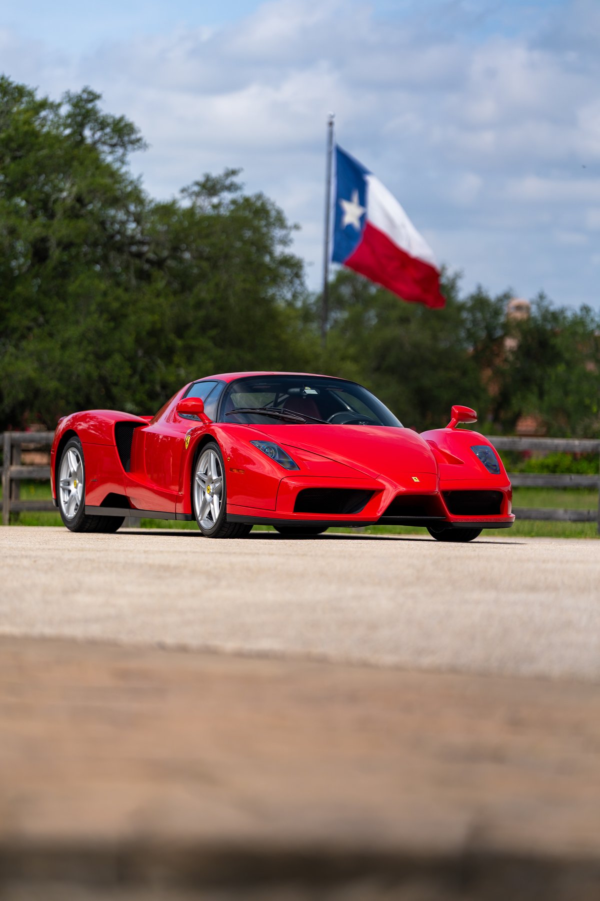A red 2003 Ferrari Enzo on silver alloy rims, with a Texas flag waving in the background.
