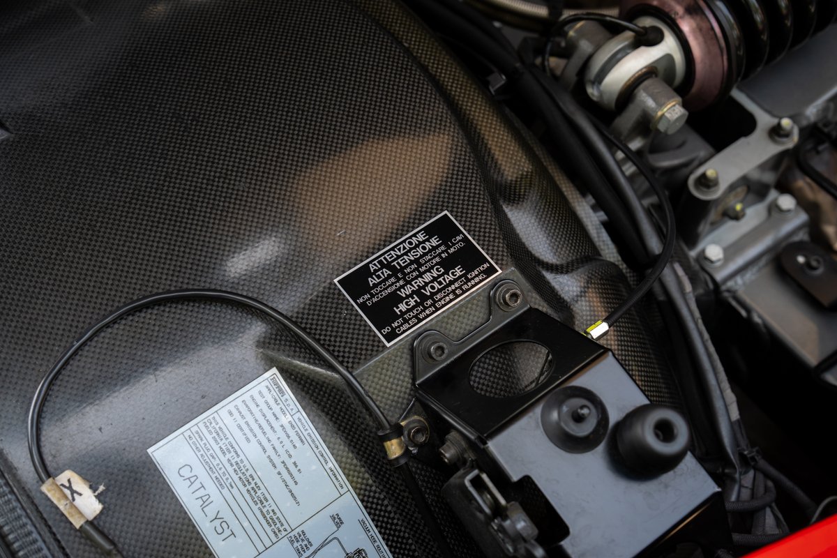 Close-up of a 2003 Ferrari Enzo engine bay, featuring carbon fiber weave and warning labels.
