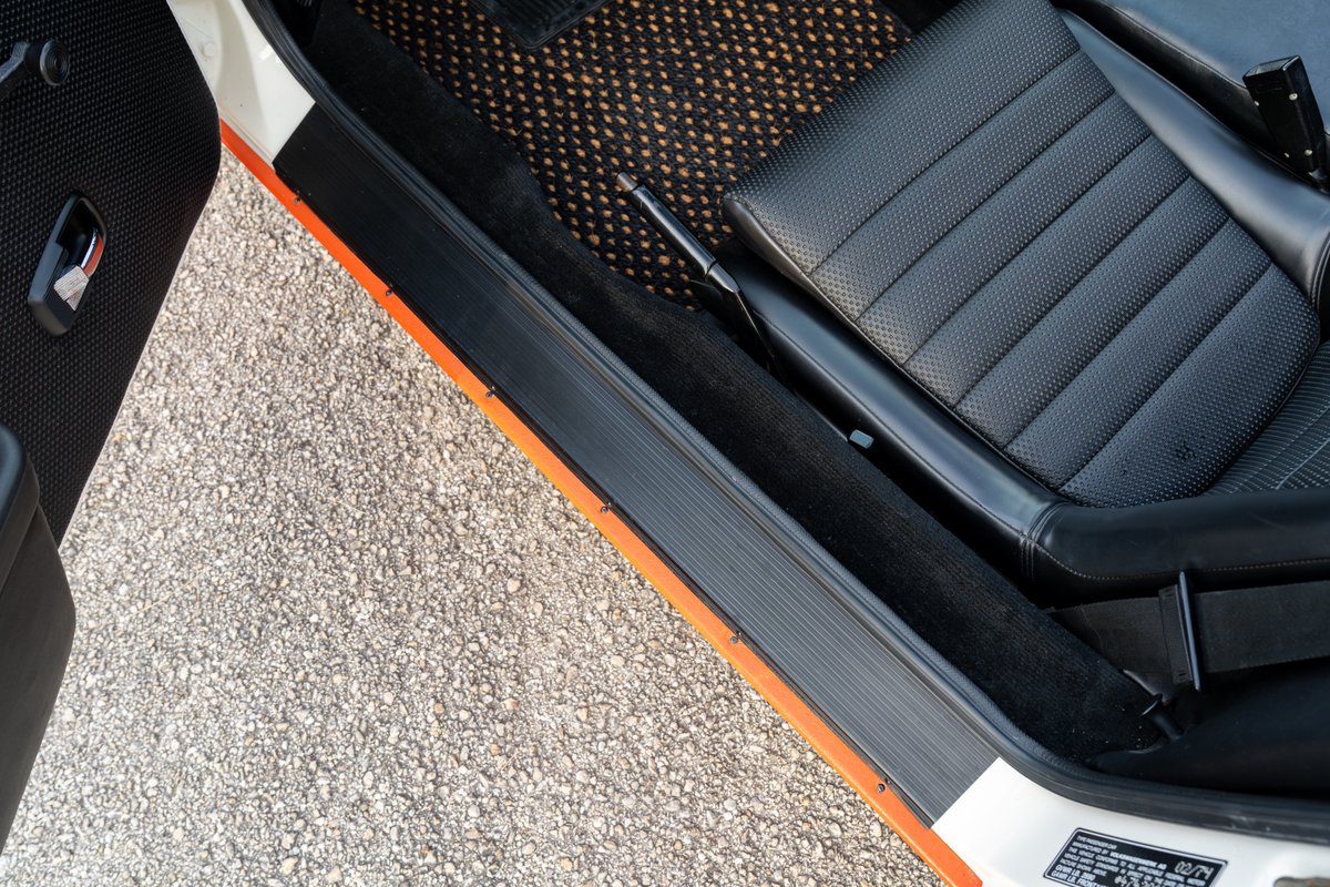 Interior view of a white 1974 Porsche 914 with orange trim, black perforated seats, and a speckled floor mat.
