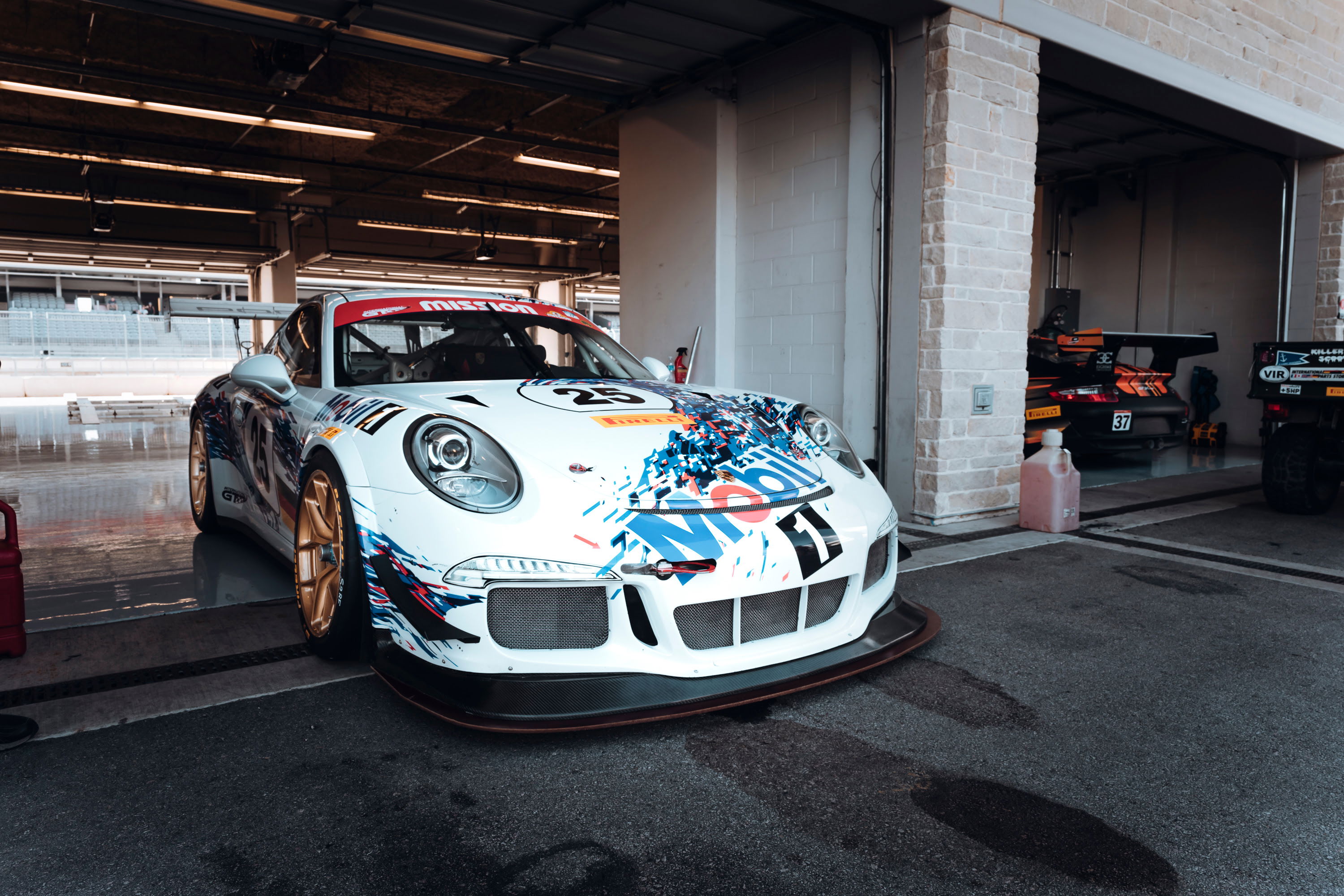 White Porsche 991 GT3 Cup race car #25 with multi-color livery, gold rims, and carbon aero parked in COTA pit lane.