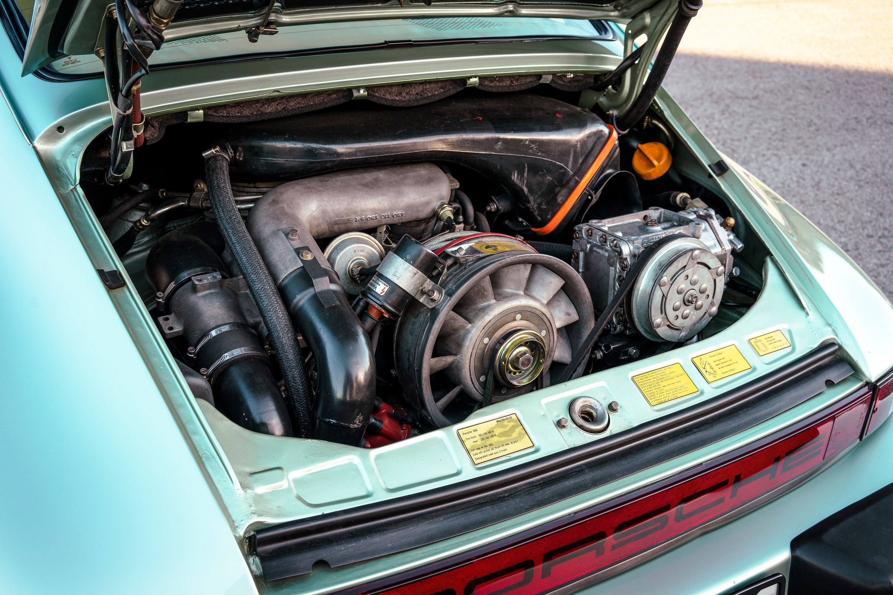 Detailed view of a 1976 Ice Metallic Green Porsche 930's rear engine bay with its air-cooled flat-six, cooling fan, and belts.