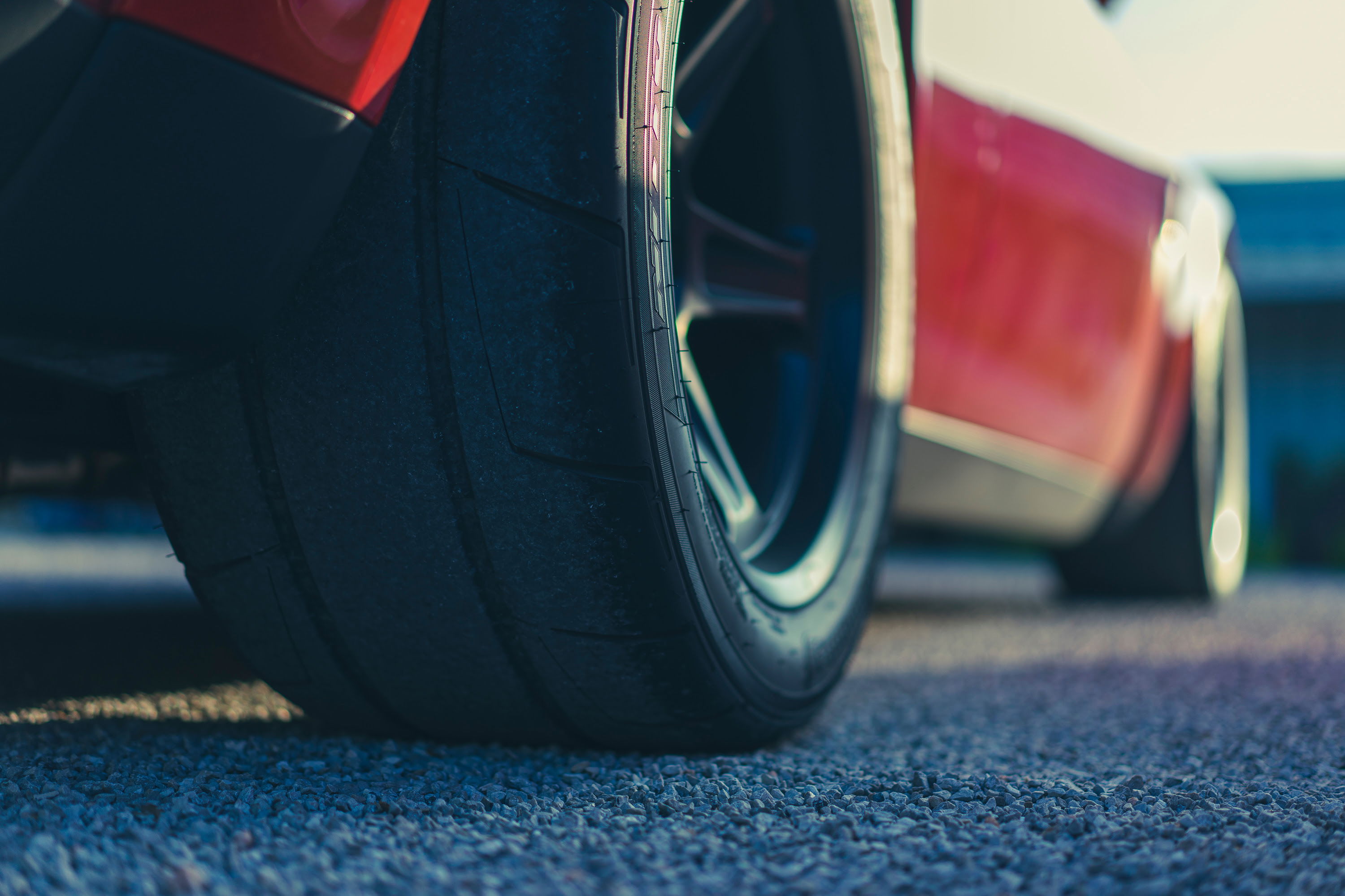 Red performance car shows off its wide, semi-slick rear tire and dark multi-spoke rim, parked on gravel.