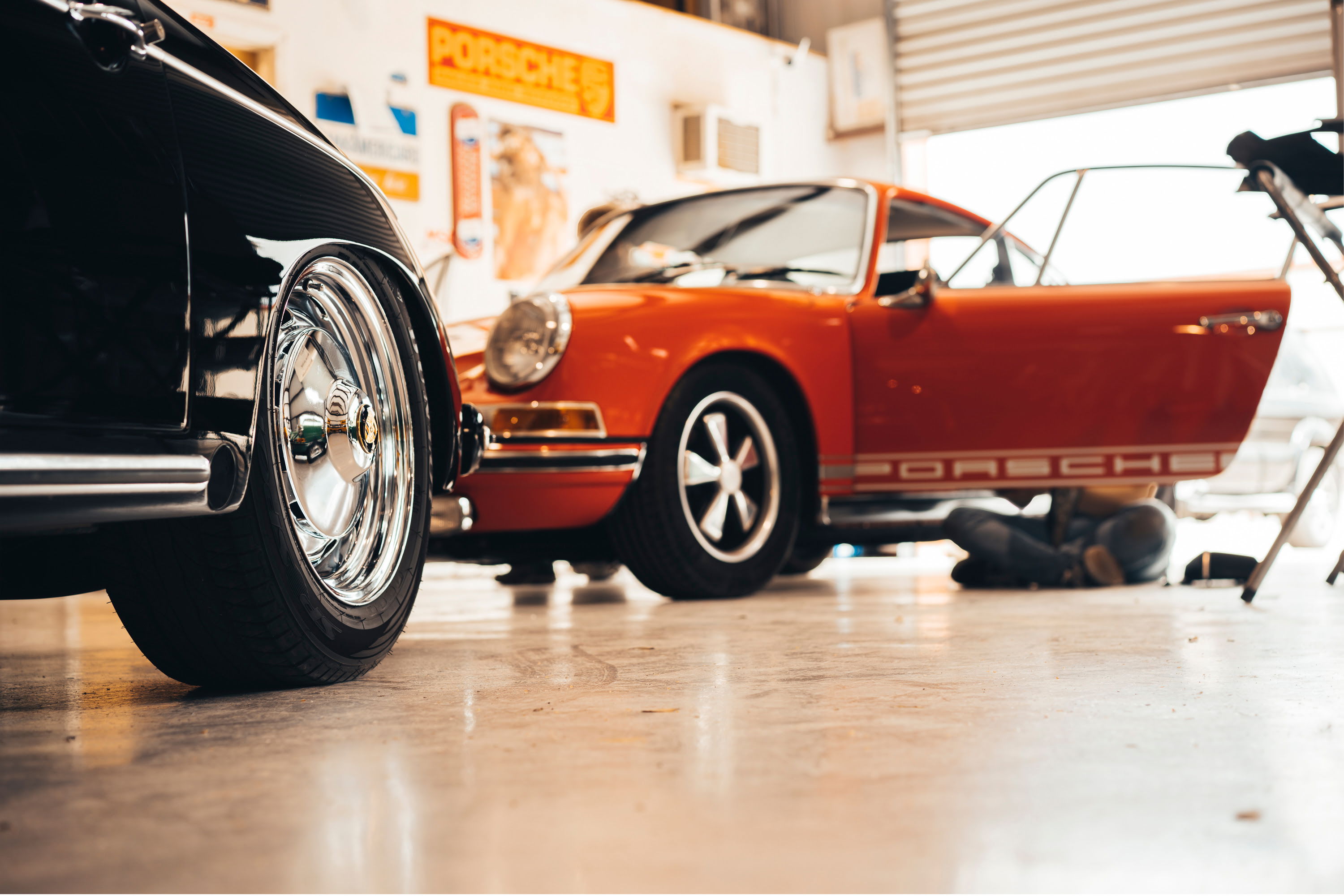 Black classic Porsche with polished chrome dish rims & side trim. Orange Porsche 911 with Fuchs wheels & 'PORSCHE' livery in shop.
