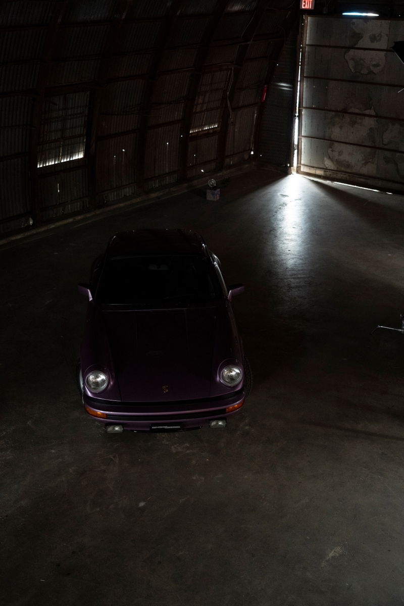 Purple Porsche 911 G-series coupe. Overhead shot showcasing classic round headlights & black bumper trim in dark industrial space.