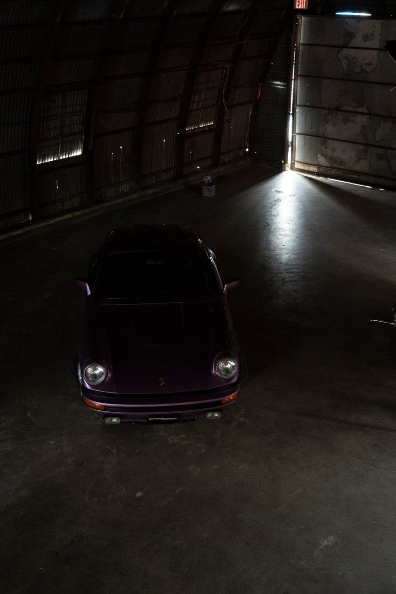 Purple Porsche 911 G-series coupe. Overhead shot showcasing classic round headlights & black bumper trim in dark industrial space.