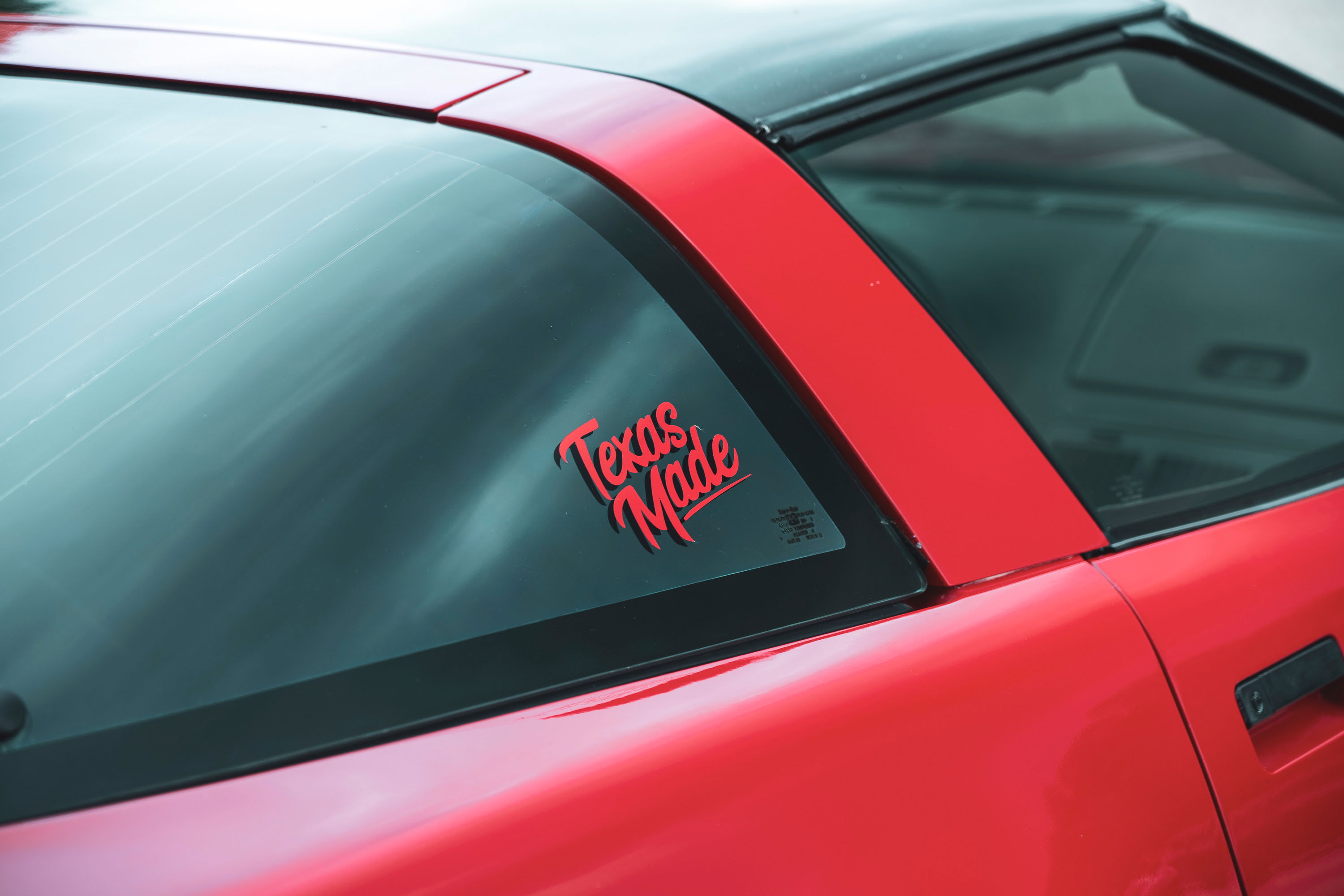 Vintage red C4 Corvette rear three-quarter view with "Texas Made" decal on dark rear window, black targa top, and sleek body lines.