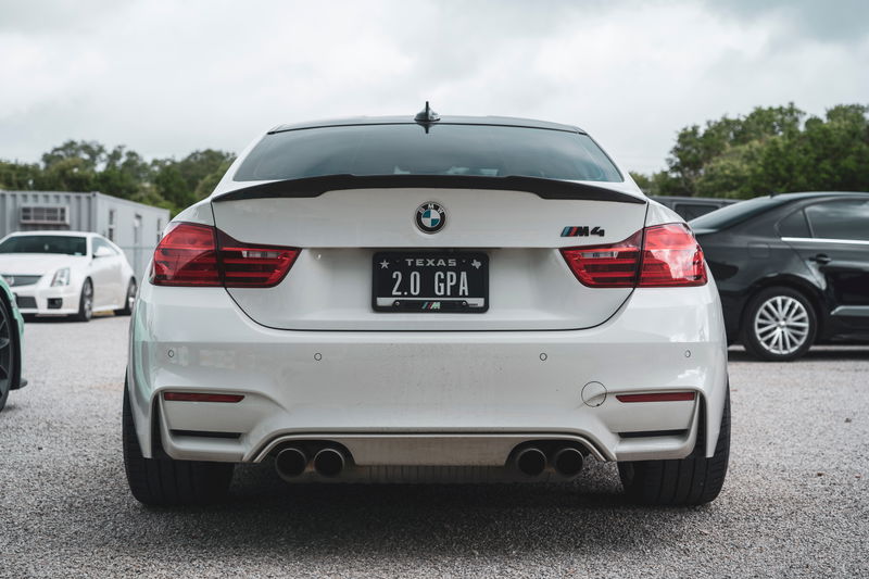 White BMW M4 (F82) rear with carbon fiber trunk spoiler, quad exhausts, and a Texas "2.0 GPA" license plate.