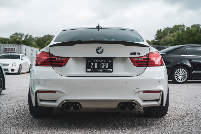 White BMW M4 (F82) rear with carbon fiber trunk spoiler, quad exhausts, and a Texas "2.0 GPA" license plate.