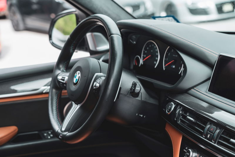 BMW X6 M (F86) interior: M leather steering wheel, carbon fiber dashboard trim, saddle brown leather accents, and red contrast stitching.