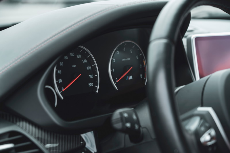 BMW M dashboard view: carbon fiber trim, red stitching, M-badged tachometer and speedometer. Modern sports car interior.