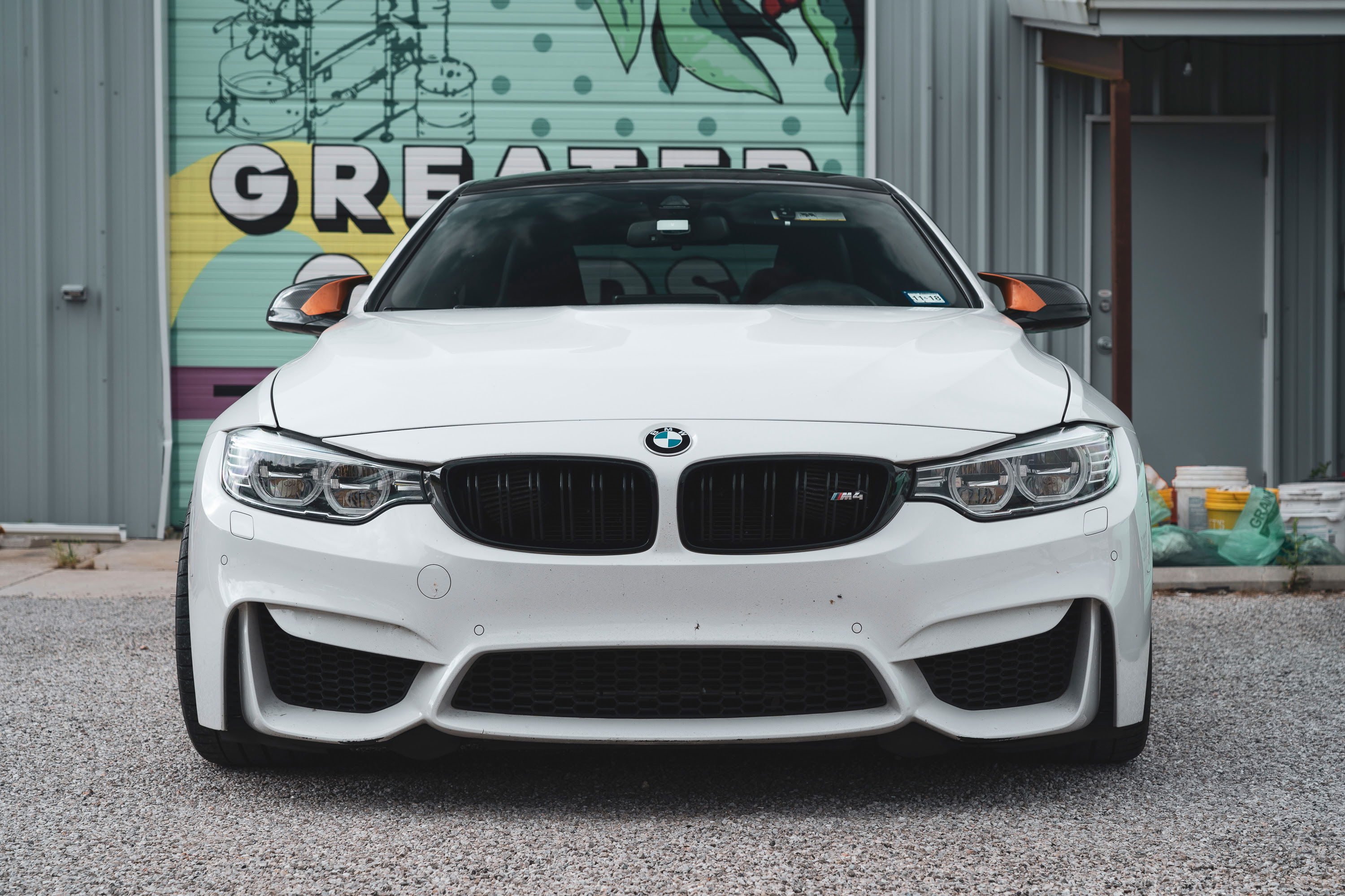 Front shot of an Alpine White F82 BMW M4 coupe, boasting black grilles, a black roof, and unique orange carbon fiber mirror trim.