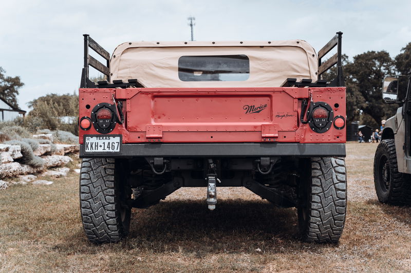 Texas-plated red Hummer H1 rear, tan canvas soft top, "Merica" tailgate, LED taillights, rugged off-road tires.