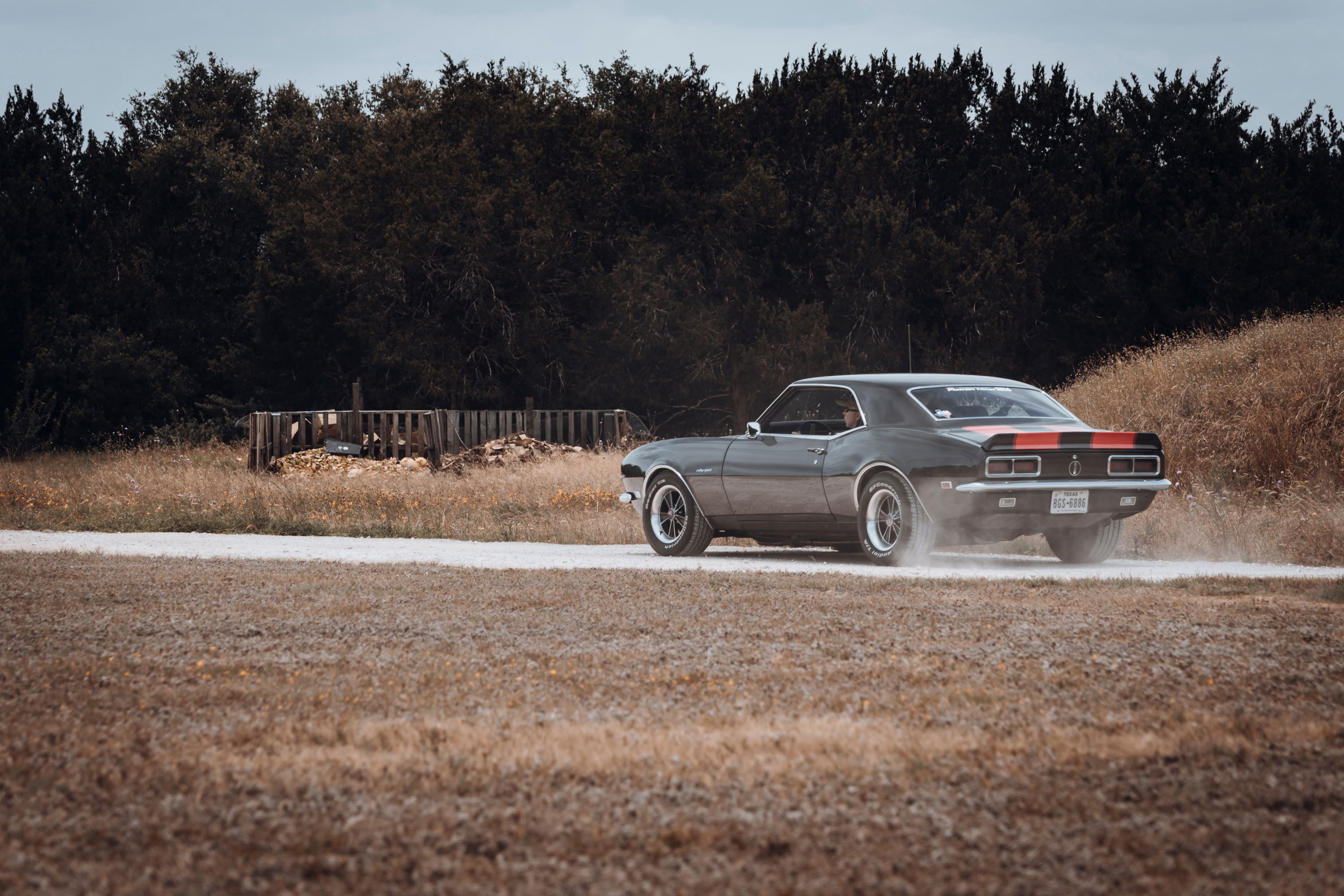 1968 Chevrolet Camaro with gray paint, red racing stripes, chrome trim, and custom multi-spoke rims kicking up dust on a dirt road.