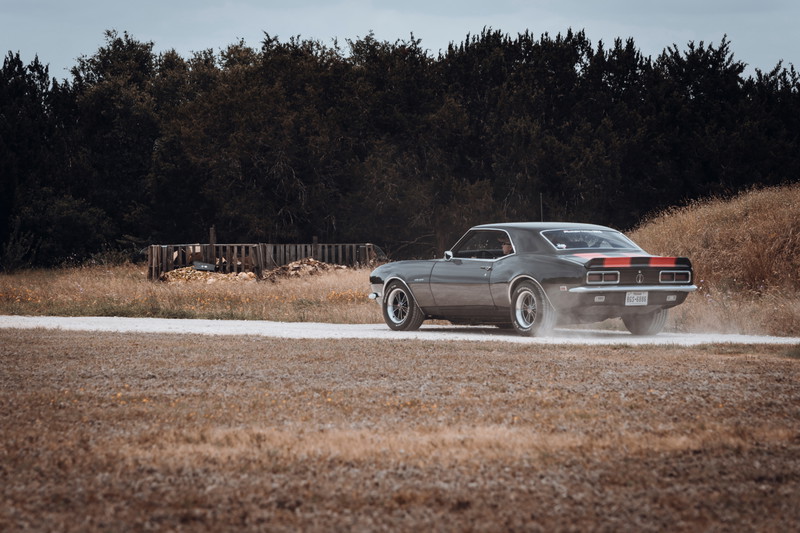 1968 Chevrolet Camaro with gray paint, red racing stripes, chrome trim, and custom multi-spoke rims kicking up dust on a dirt road.