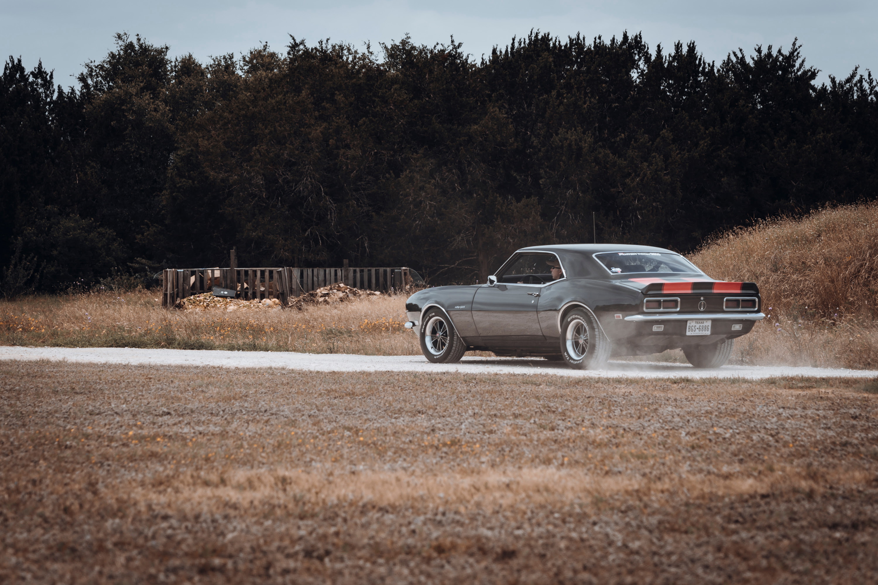 1968 Chevrolet Camaro with gray paint, red racing stripes, chrome trim, and custom multi-spoke rims kicking up dust on a dirt road.