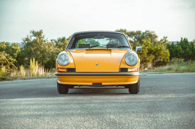 Front view of a 1973 Signal Yellow Porsche 911S, showcasing iconic round headlights, black bumper trim, and Fuchs wheels.